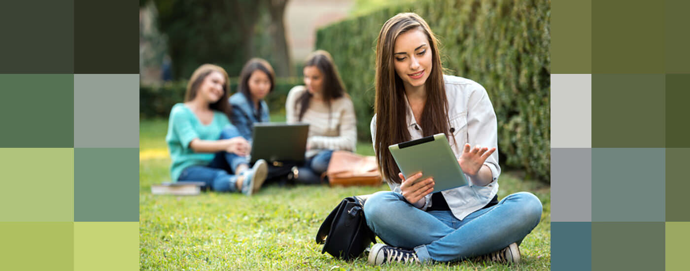 a student sitting on the grass, using a tablet. students in the background are shown starting at a laptop.