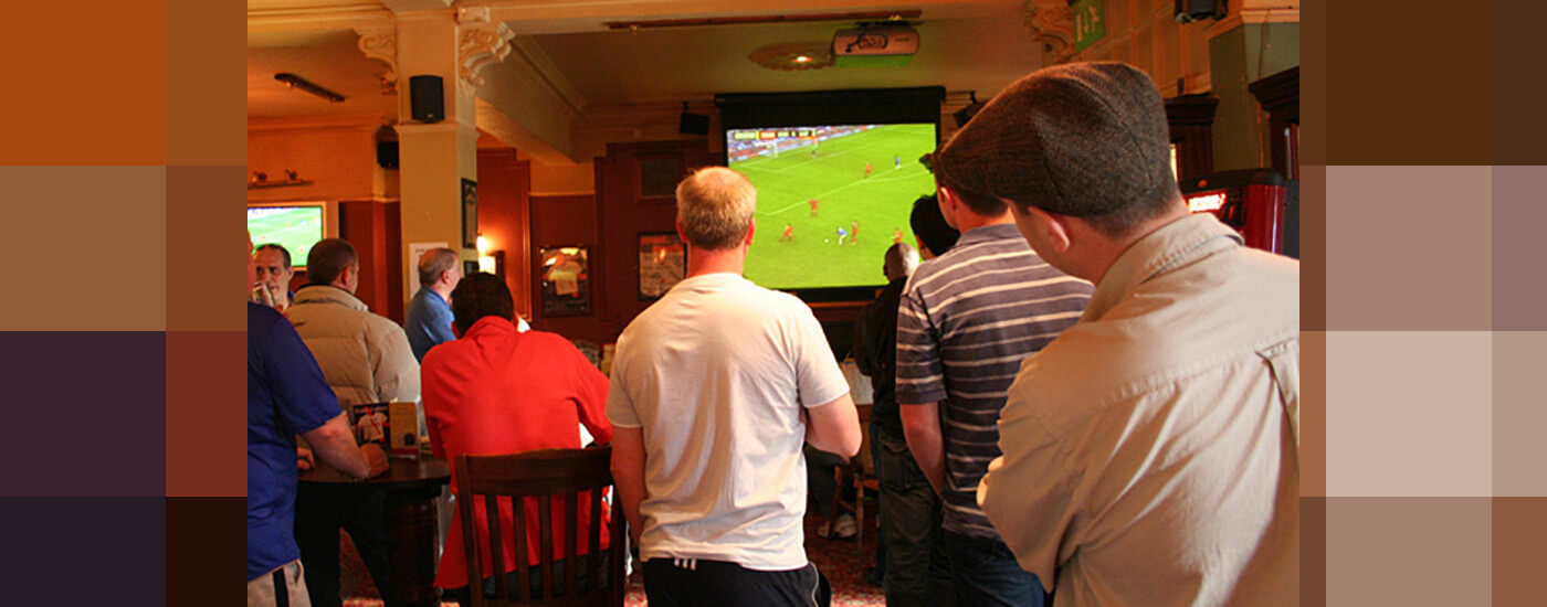 The backs of men as they look onto a screen playing a soccer game at a pub