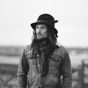 Ian Treherne poses for the photo while wearing a hat, jean jacket, and scarf. The photo is in black and white.