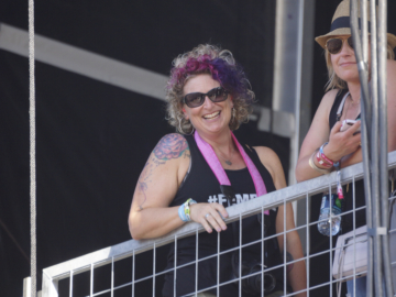 Cat Brewer poses up on a balcony and smiles down towards the camera. She wears black sunglasses, a tank top, and has pink and purple curly hair.
