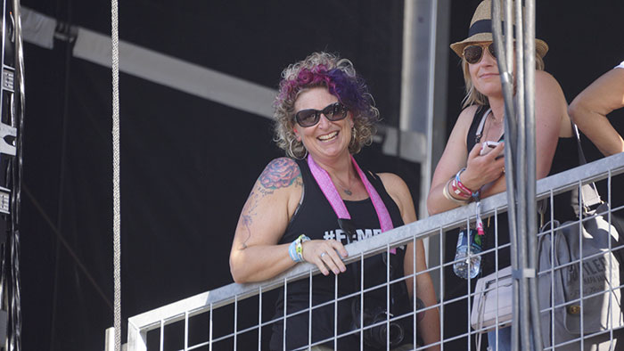 Cat Brewer poses up on a balcony and smiles down towards the camera. She wears black sunglasses, a tank top, and has pink and purple curly hair.