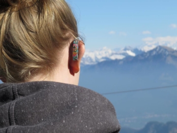 E faces the Swiss Alps. She is wearing a hearing aid with a colorful pattern.