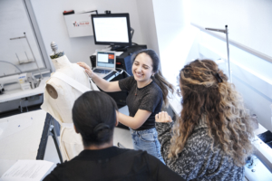 Camila smiles while showing two other women her designs on a mannequin 