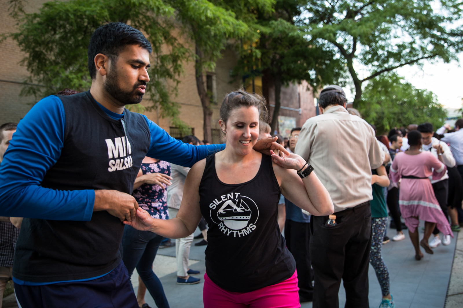 Kerry dances salsa in a park with a man. She is wearing a black silent movements tank top and pink pants.