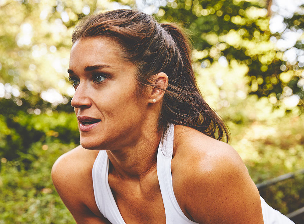 Rebecca wears a white athletic tank top with her hair in a high ponytail while posing in front of vibrant green trees.