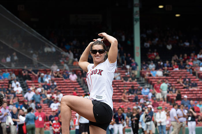 Rebecca Alexander wears a Red Sox jersey and has a big grin as she throws the first pitch at a Boston Red Sox baseball game.