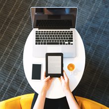 Someone in front of a laptop, a smartphone, and a cup of coffee, reading on a tablet