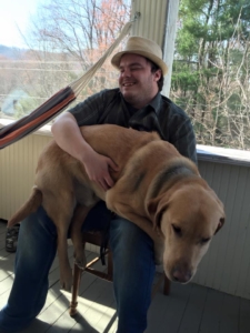 Josh Pearson sits on a chair outside on a porch smiling and holding his golden Lab Alpha