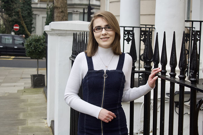 Elin Williams smiles wearing a white ribbed turtleneck under a dark denim dress. She stands in the street of her down against a black iron fence.