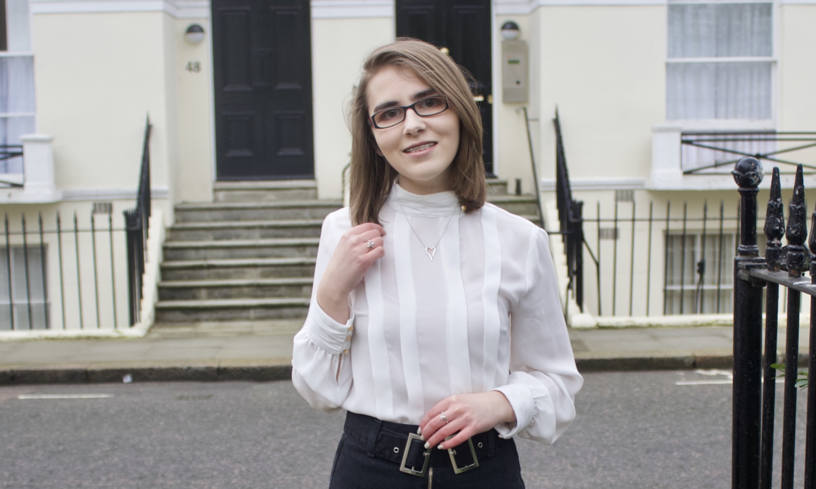 Elin wears a white blouse and black pants. She stands in front of a building smiling and touching her hair.