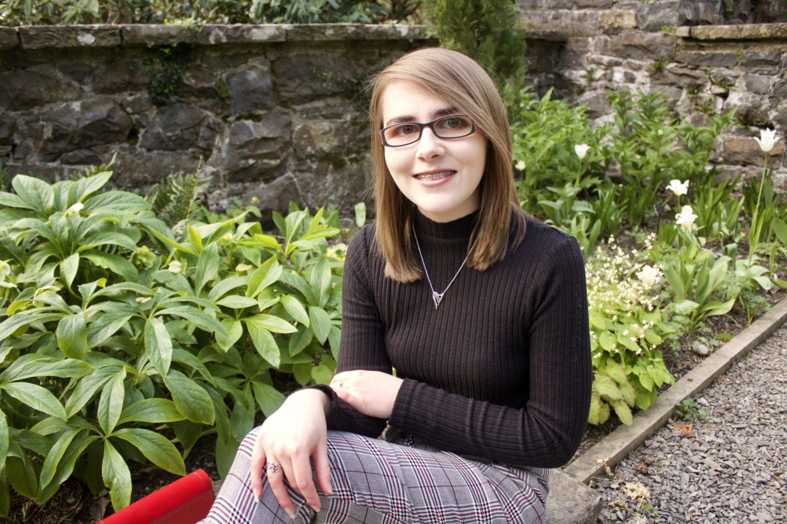 Elin sits with her legs crossed in front of a stone wall and greenery. She wears black glasses, plaid pants, and a black turtleneck.