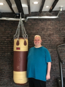 Gus poses next to a punching bag in a gym while smiling and wearing a blue shirt and black pants