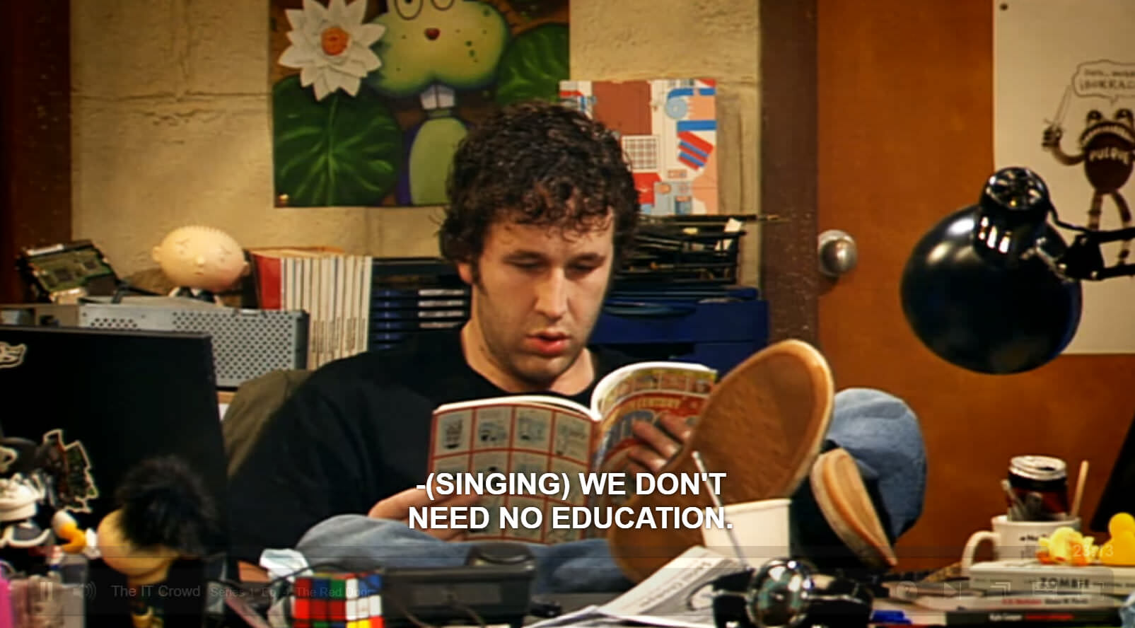 A screenshot from the IT Crowd in which Roy is seated at his desk reading a magazine. The caption reads, (Singing) We don't need no education. The word Singing is in brackets.