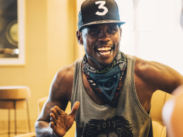 a man smiling in a chair mid conversation wearing a hat with a 3 on it, a bandanna around his neck, a sleeveless shirt with a lion on it, and basketball shorts