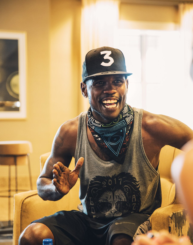 a man smiling in a chair mid conversation wearing a hat with a 3 on it, a bandanna around his neck, a sleeveless shirt with a lion on it, and basketball shorts 
