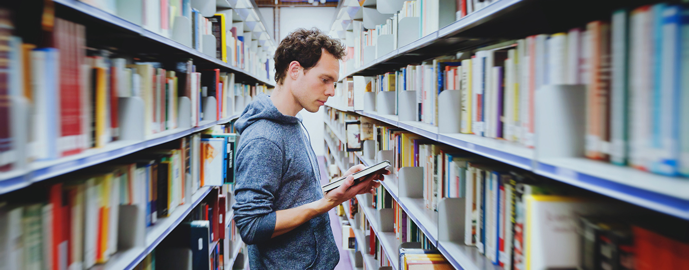 Man looking at a book in his hand in between two aisles filled with shelves of books