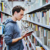 Man looking at a book in his hand in between two aisles filled with shelves of books
