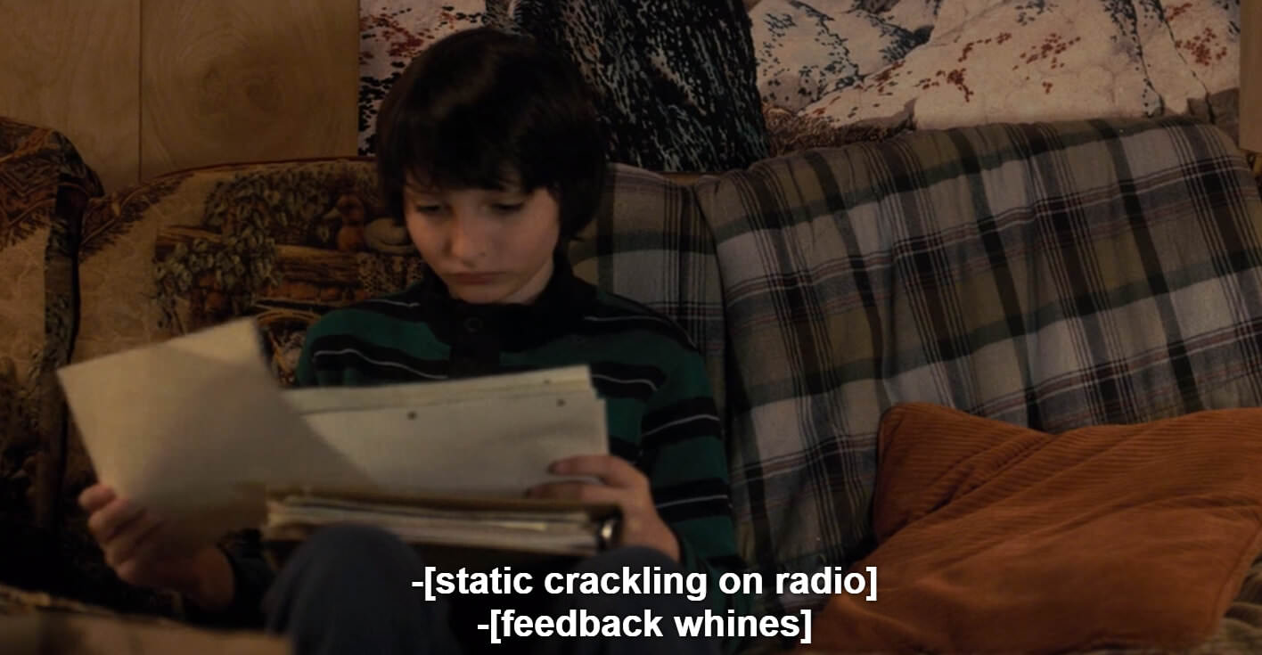 A screenshot from Stranger Things in which Mike sits on a couch shuffling papers. The two lines of captions in brackets read [static crackling on radio] and [feedback whines]